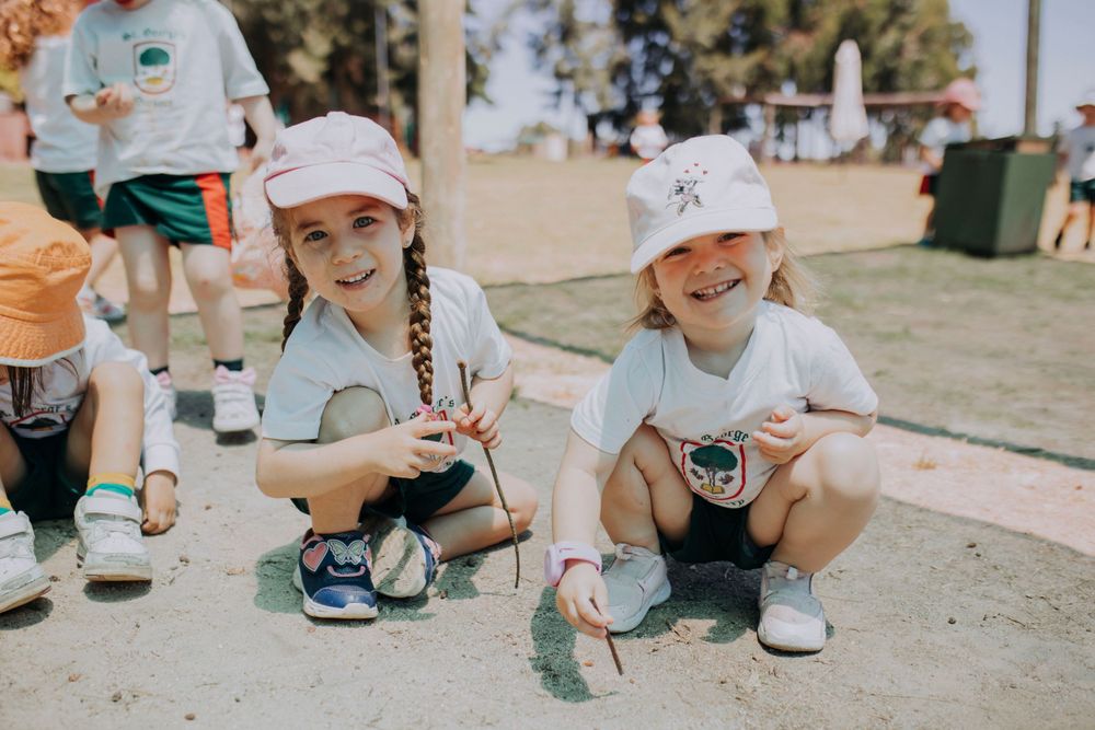 Two children in baseball caps crouch on the ground holding drinks and enjoying a sunny day outdoors.