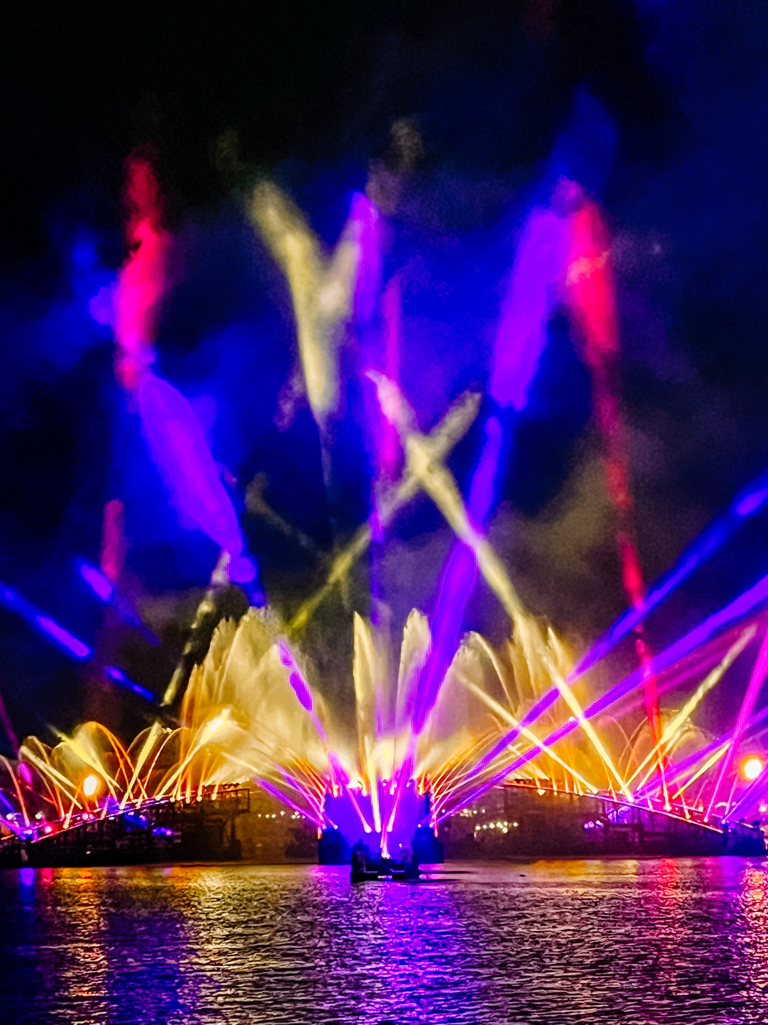 Vibrant laser light show reflects on water with blue, pink and yellow beams shooting into dark sky above illuminated fountains.