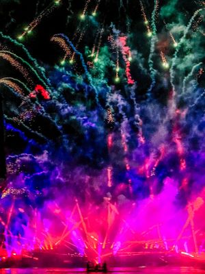 Spectacular fireworks display illuminates the night sky in vibrant pink, blue and green colors against a water show.