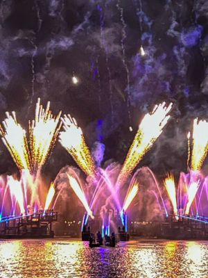 Spectacular nighttime fireworks and colorful fountains display reflected in water with vibrant blue and pink illumination.