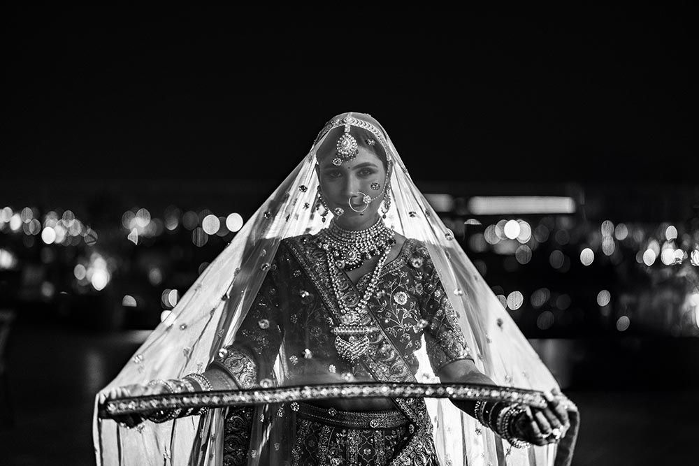 A bride wearing traditional Indian attire and veil stands against a bokeh light background at night.