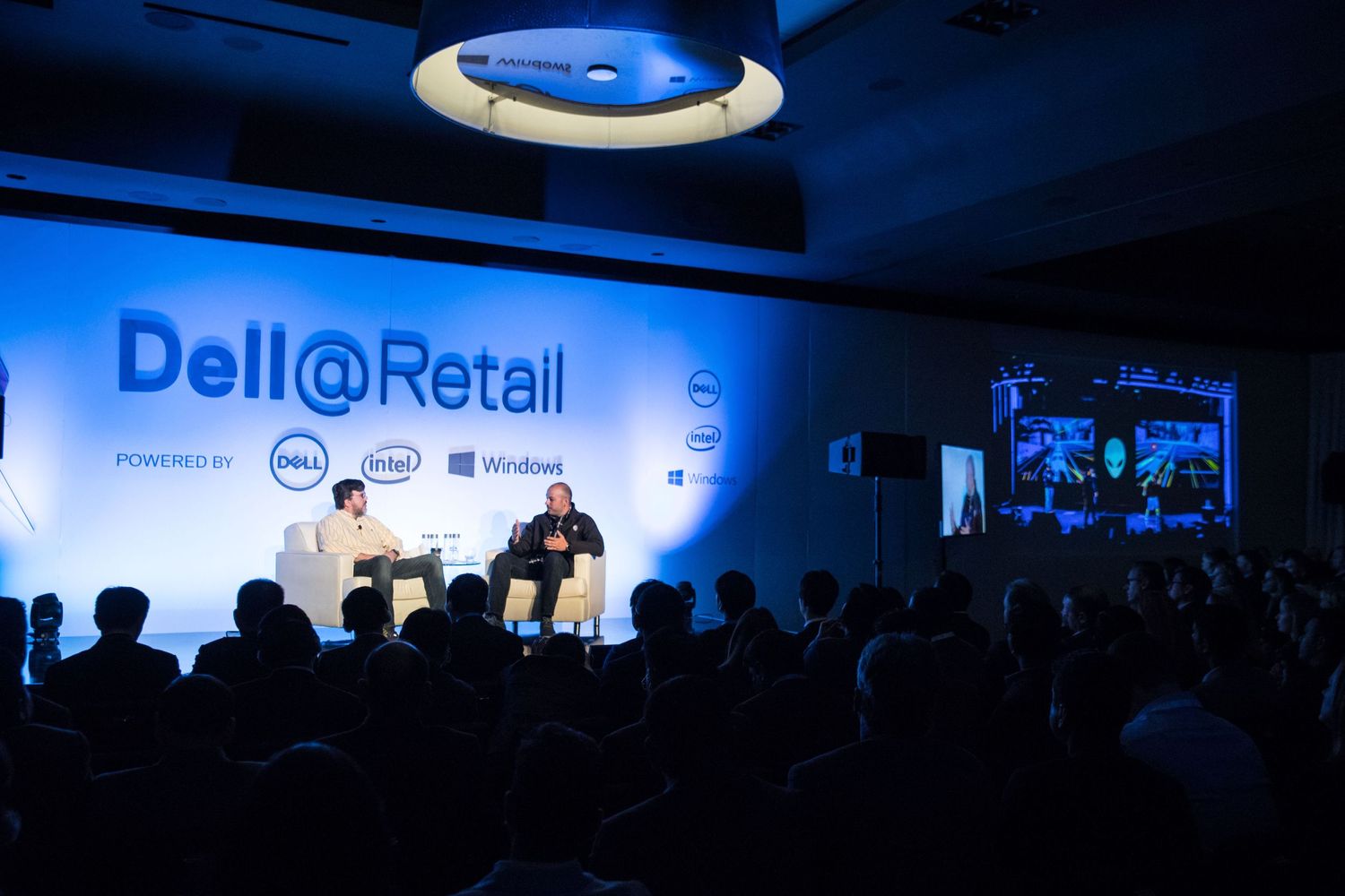 A dark auditorium with blue lighting and a Dell@Retail stage setup hosting a business conference presentation.
