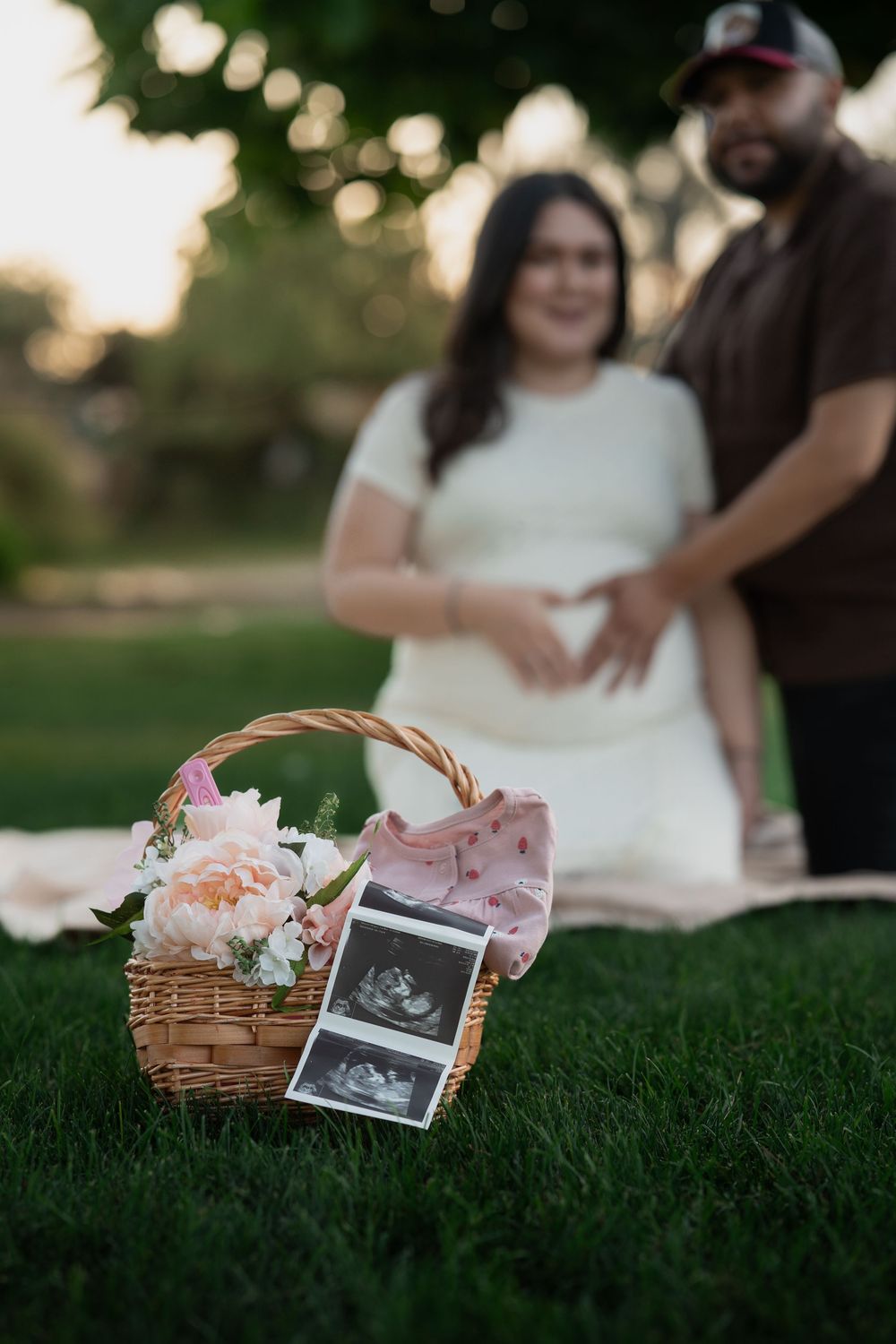 A wicker basket with baby items and ultrasound photos arranged on green grass during sunset.