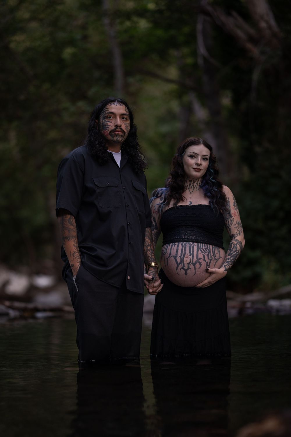 Couple in black clothing poses for dramatic maternity photos near water at dusk.