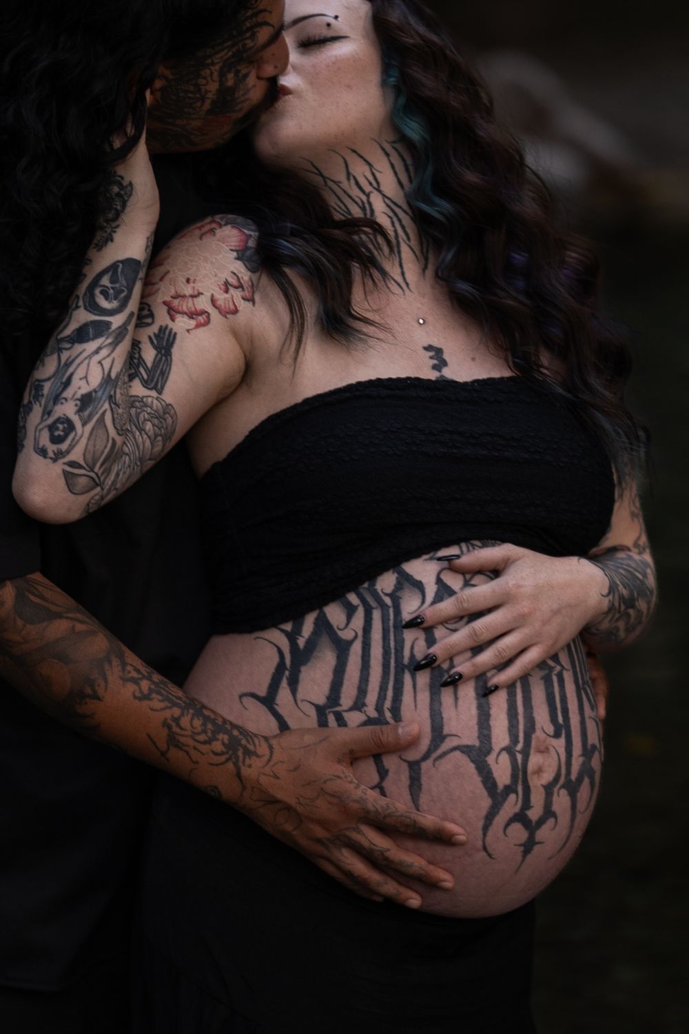 Close-up details of tattooed arms cradling a pregnant belly in dark artistic photography.