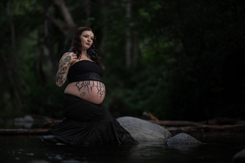 Moody maternity photoshoot by water featuring dark clothing and natural scenery.