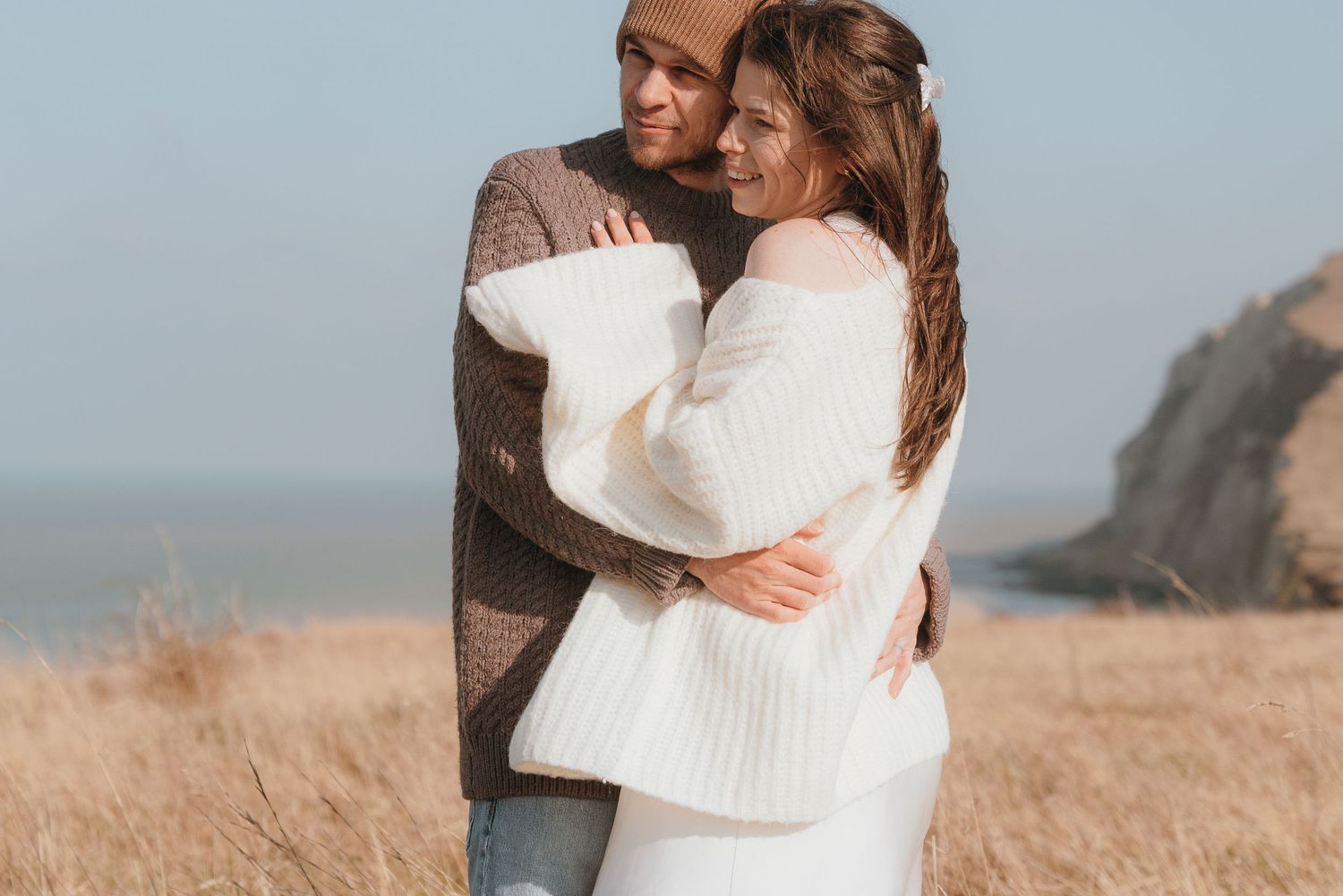 A romantic couple embraces on a coastal cliff overlooking the ocean on a sunny afternoon.