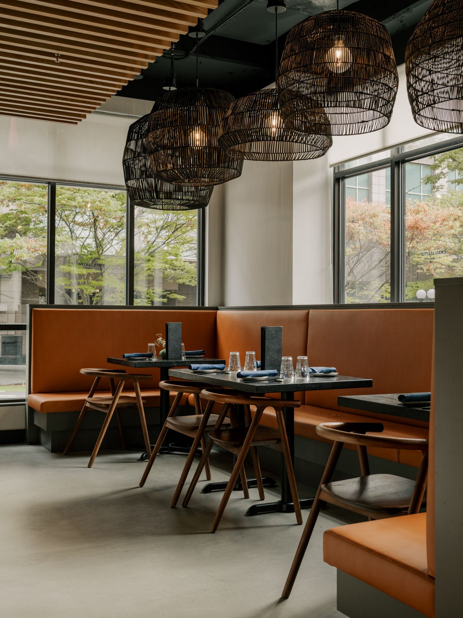 Modern restaurant interior with woven pendant lights, brown leather banquette seating, and wooden tables against large windows.