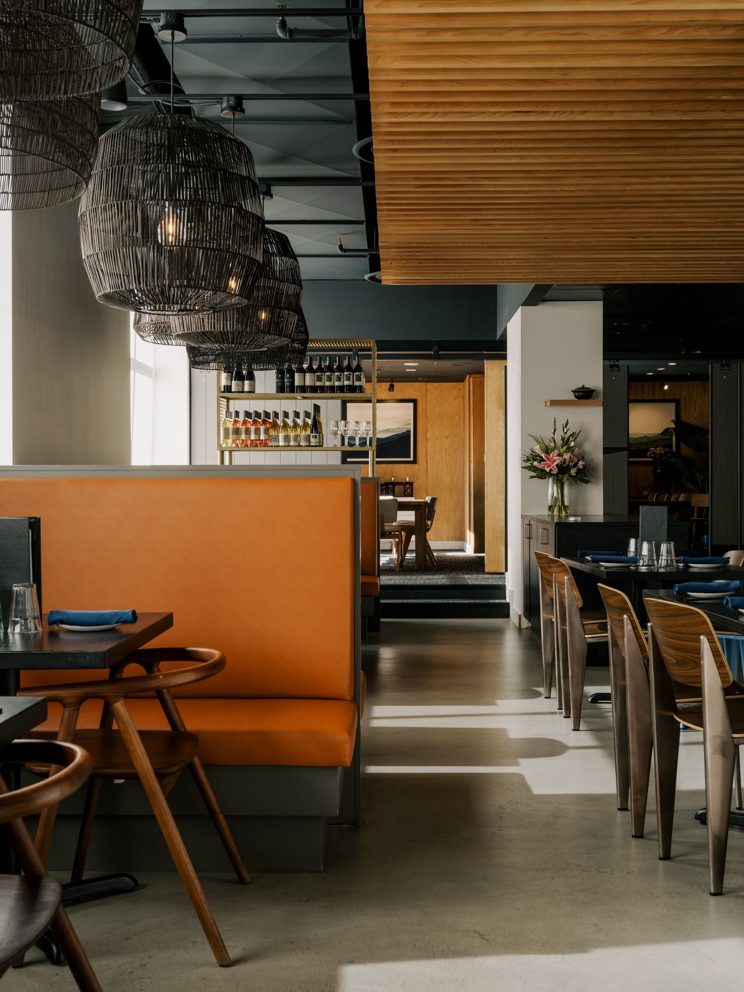 Modern restaurant interior with orange banquette seating, woven pendant lights, and wood paneled walls in warm lighting.