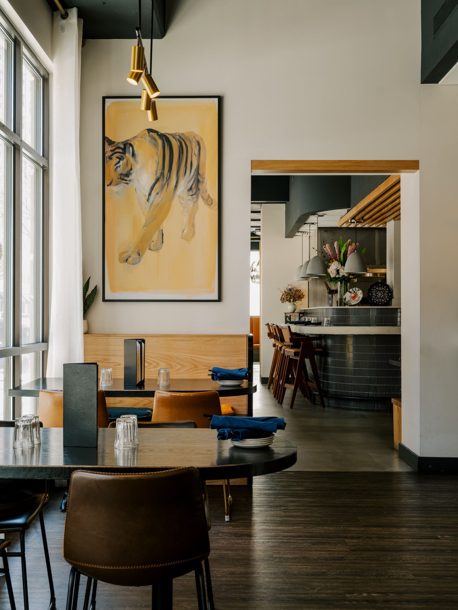 Modern restaurant interior with wooden tables, leather chairs, and Asian-inspired decor including a large tiger painting on yellow wall.