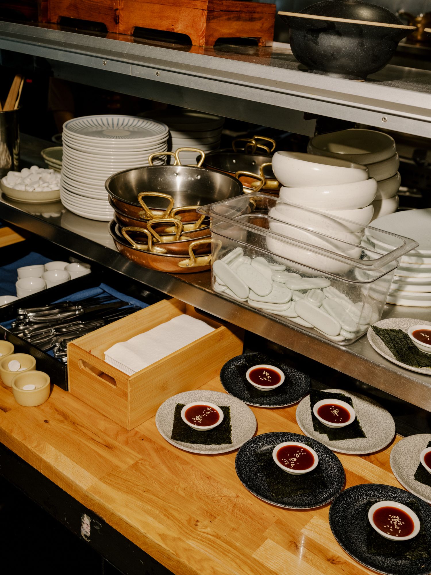 Restaurant kitchen storage shelves with stacked white plates, metal cookware, and plated desserts arranged on wooden counter.
