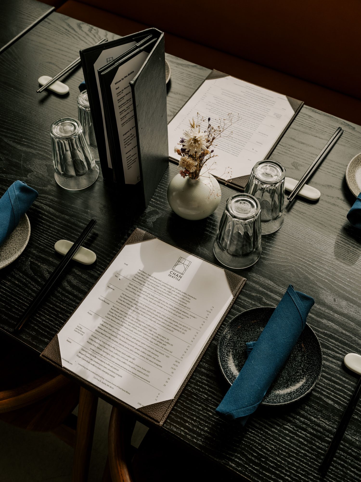 Restaurant table setting with menus, drinking glasses, blue napkins, and a small vase of dried flowers on dark wooden surface.
