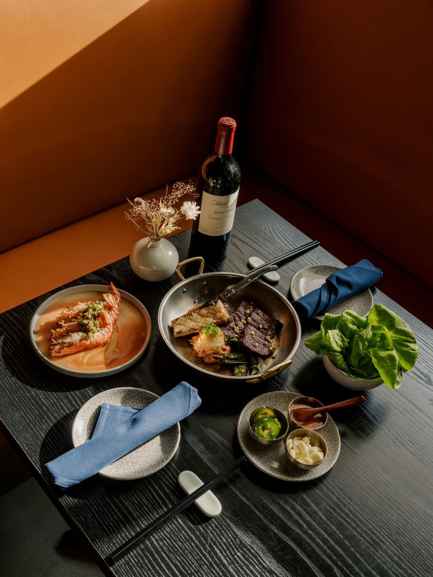 Elegant table setting with wine, steak in skillet, bowls of side dishes, and blue napkins on dark wooden surface in moody lighting.