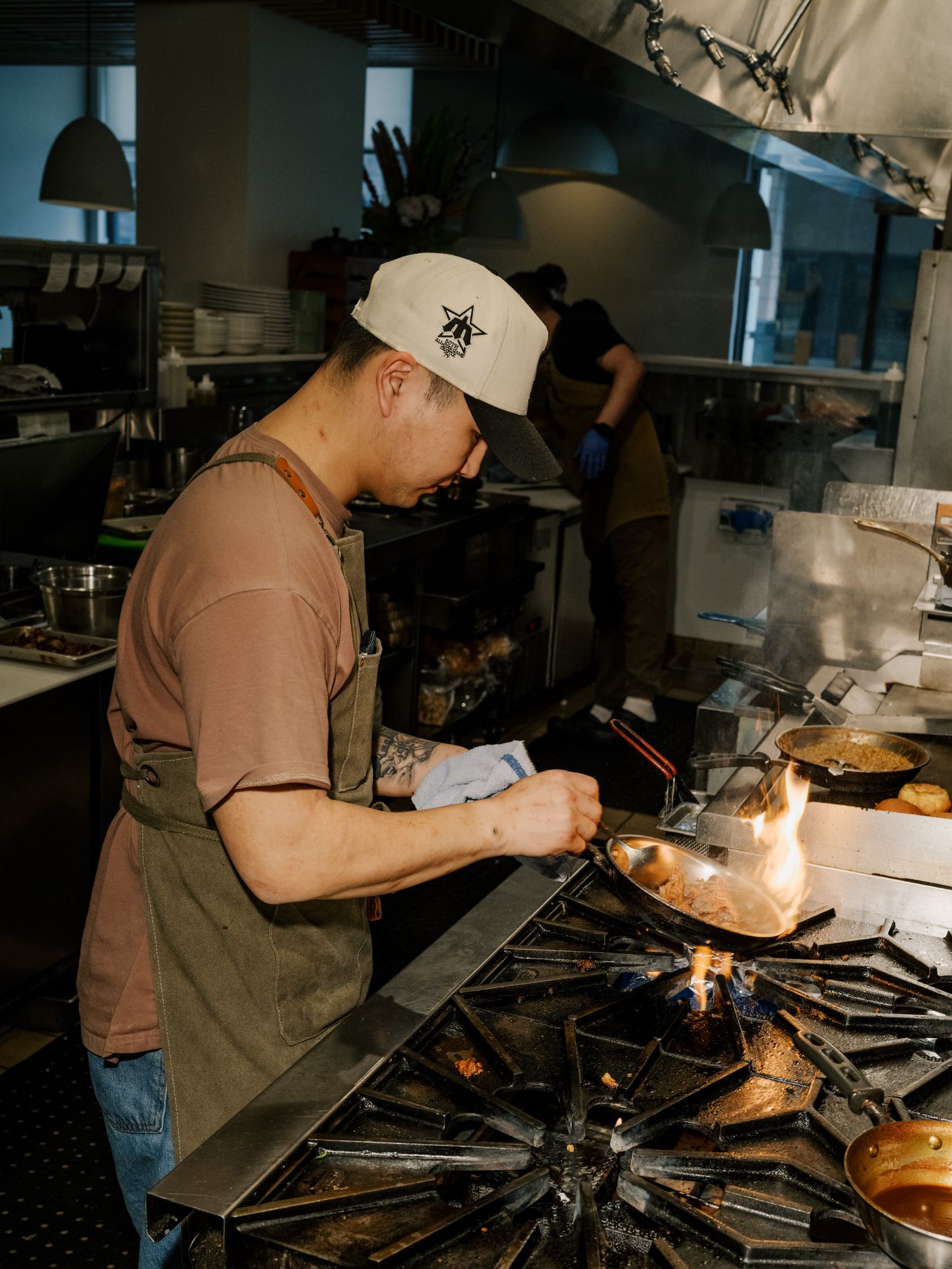 Chef in brown shirt and baseball cap cooking at busy stovetop while checking order ticket in commercial kitchen.