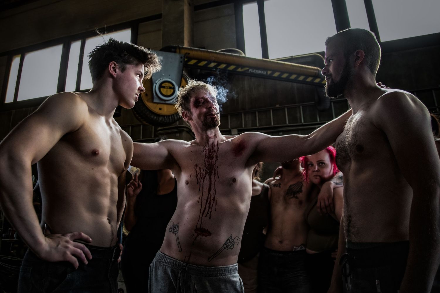 Three shirtless figures in an industrial setting with dramatic lighting.