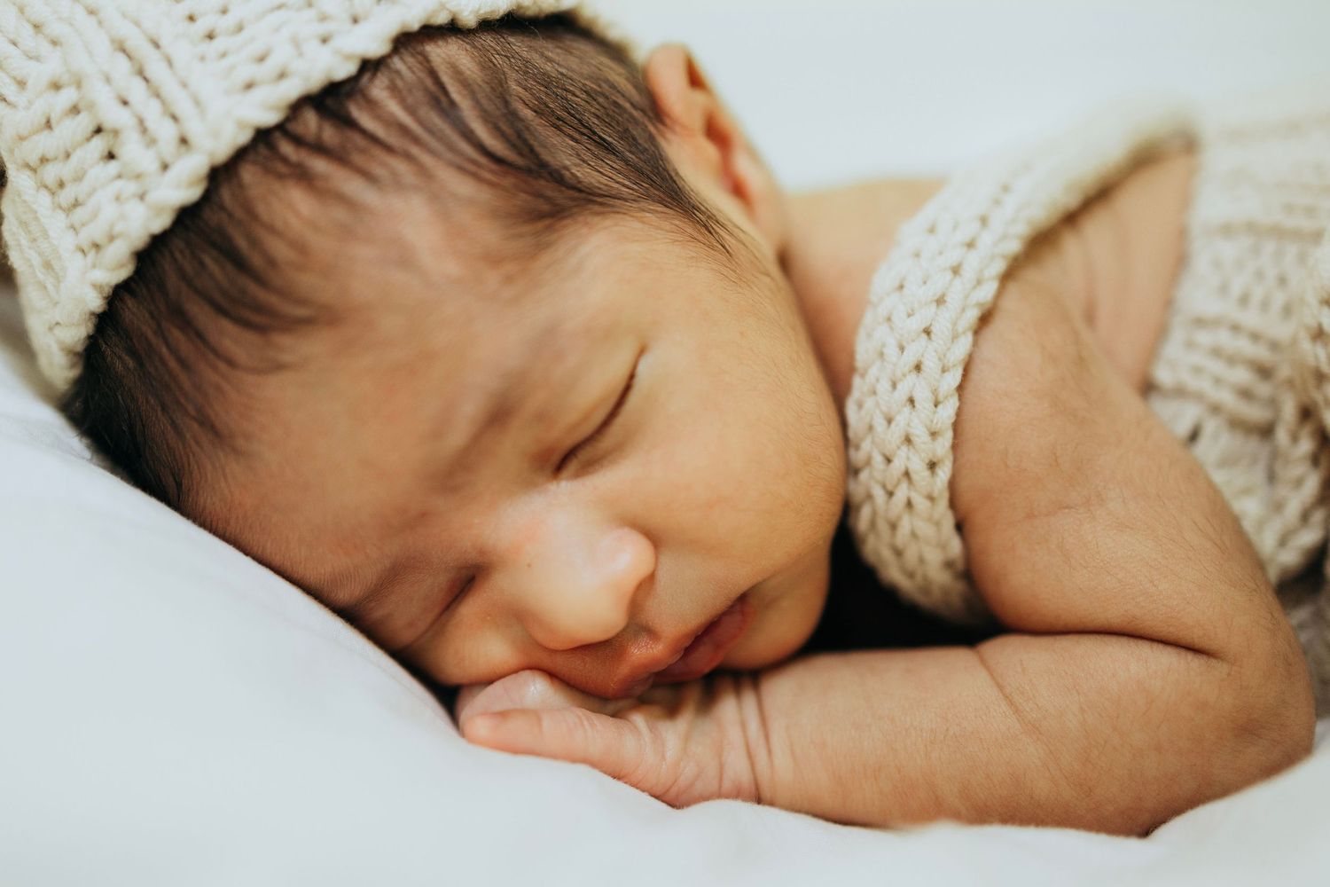 Sleeping newborn wearing knit hat rests peacefully on white bedding.