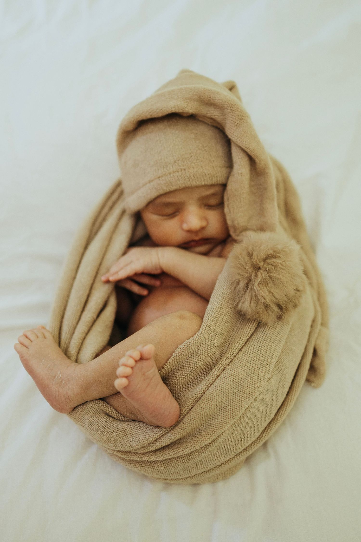 Sleeping baby wrapped in tan blanket with pom pom detail on white background.