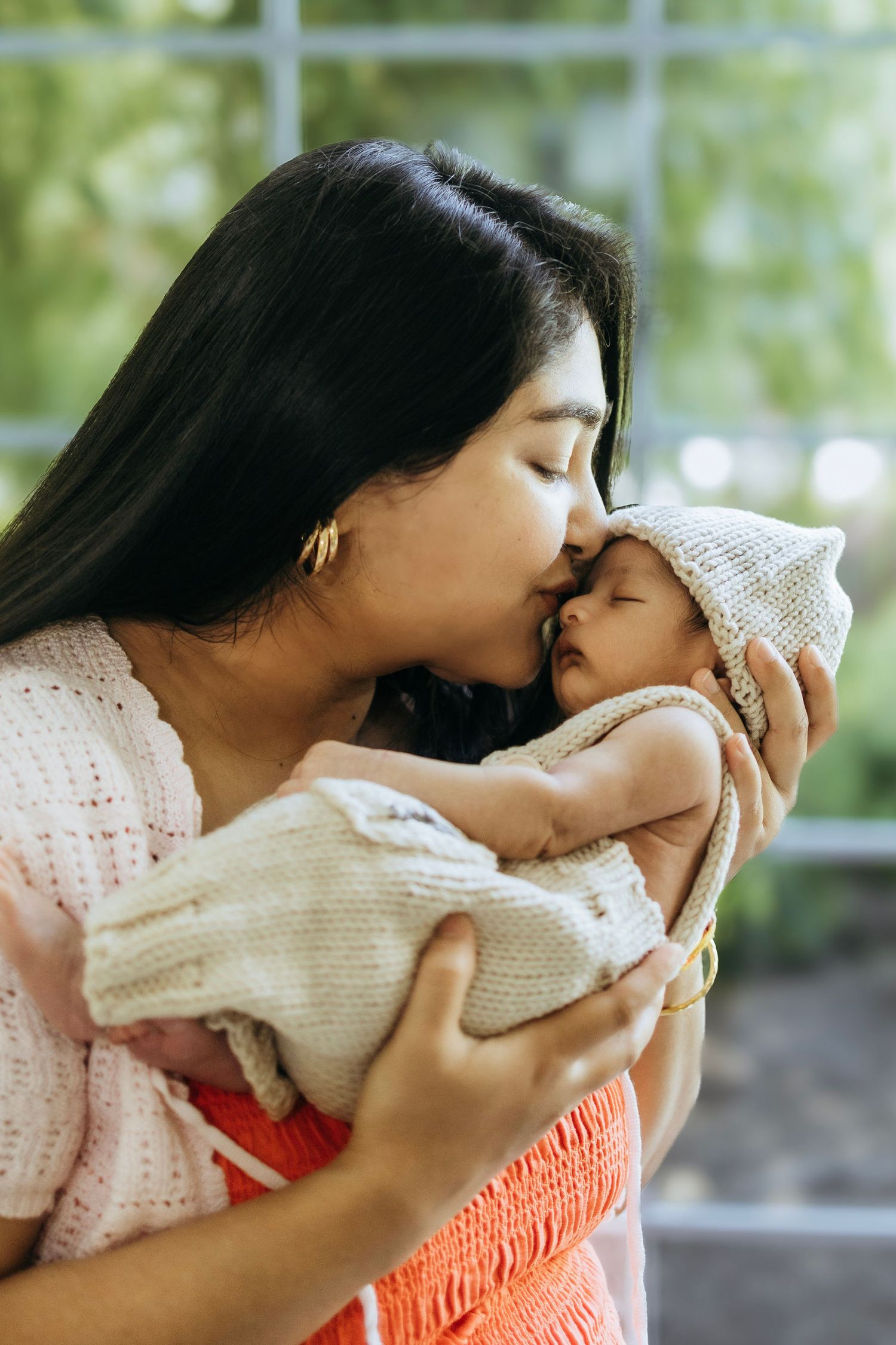 Tender moment between mother and baby wrapped in knit blanket by window.