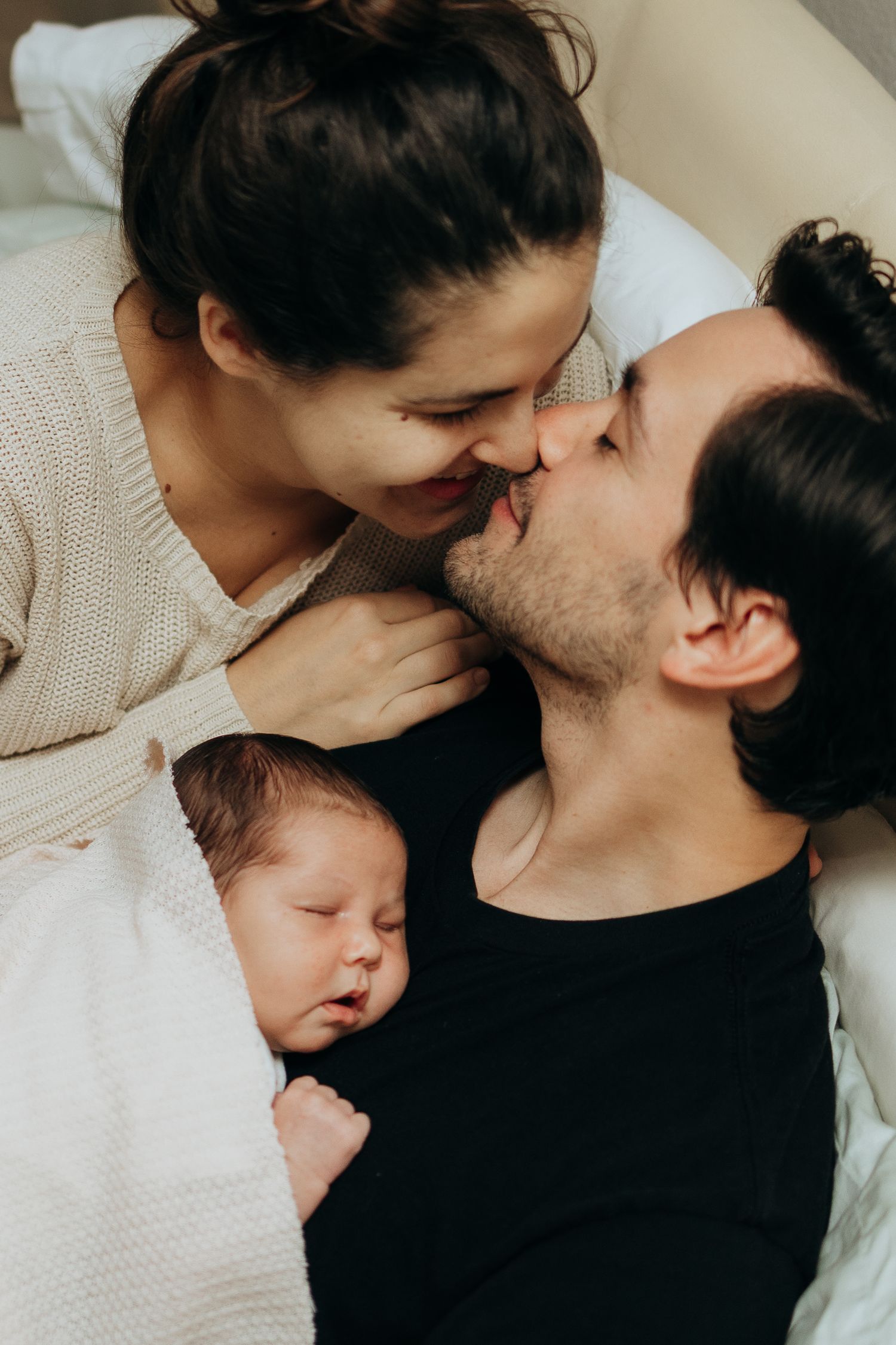Family cuddle session with newborn baby in soft indoor lighting.