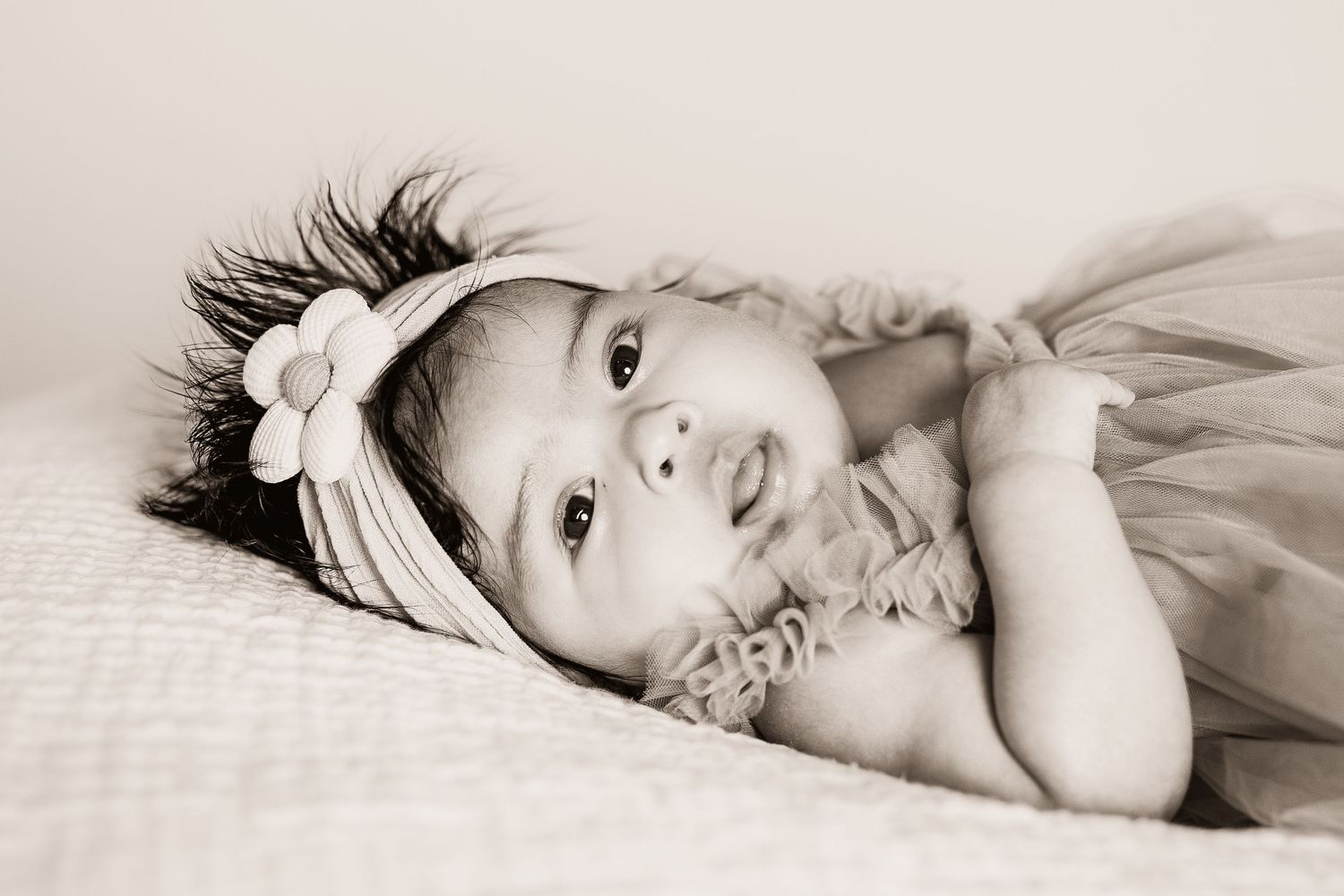 Black and white photo of a cute newborn baby girl wearing a flower headband lying on a soft blanket.