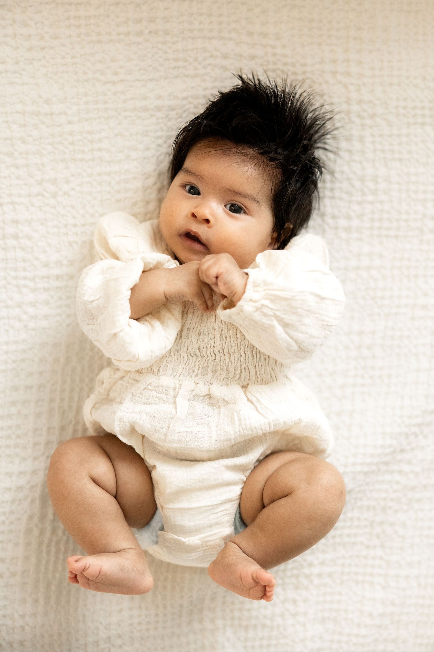 Adorable baby with dark spiky hair lying on white blanket, wearing cream outfit, looking up curiously.
