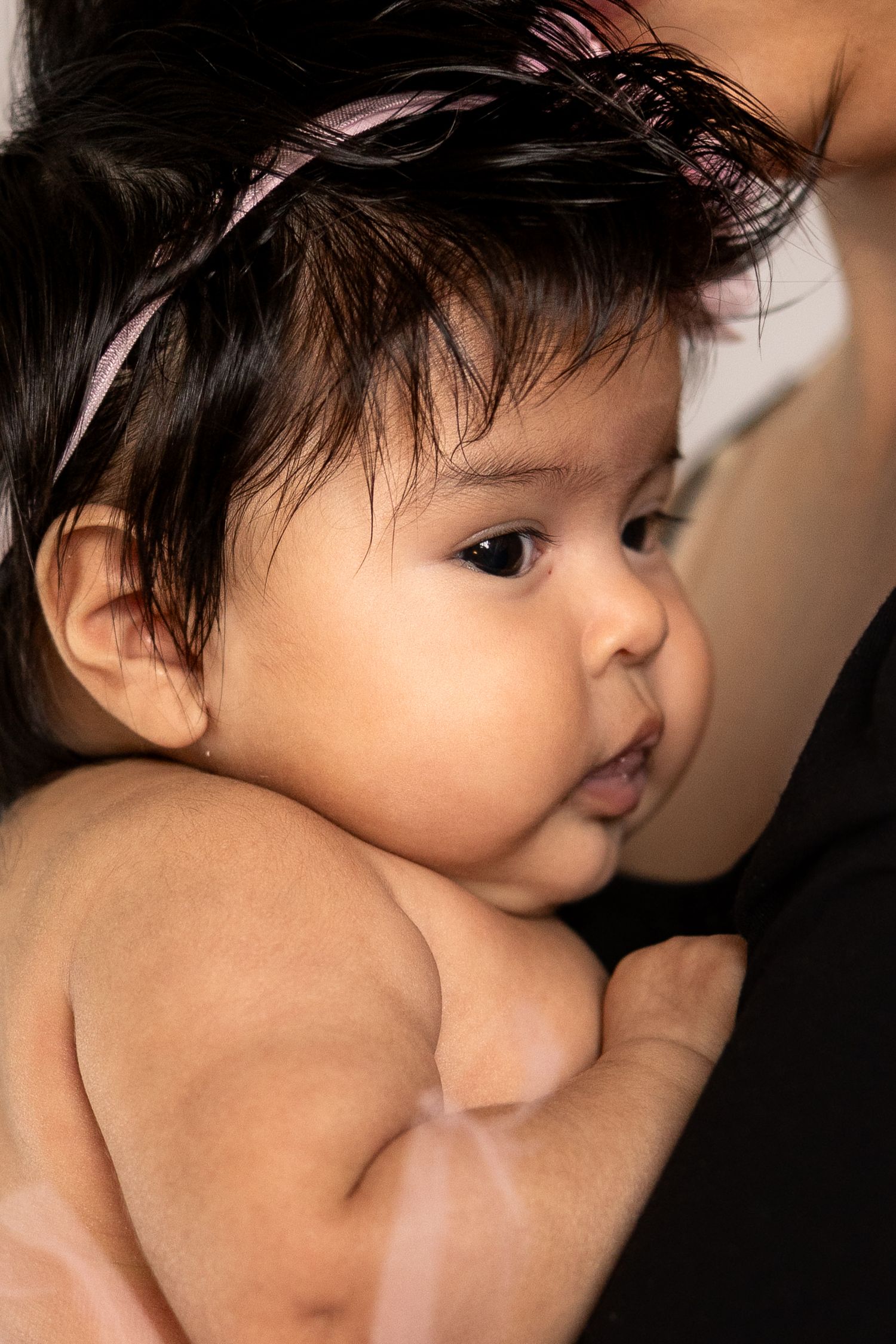 A chubby dark-haired baby girl with a pink headband being held by her mother, looking alert and curious.