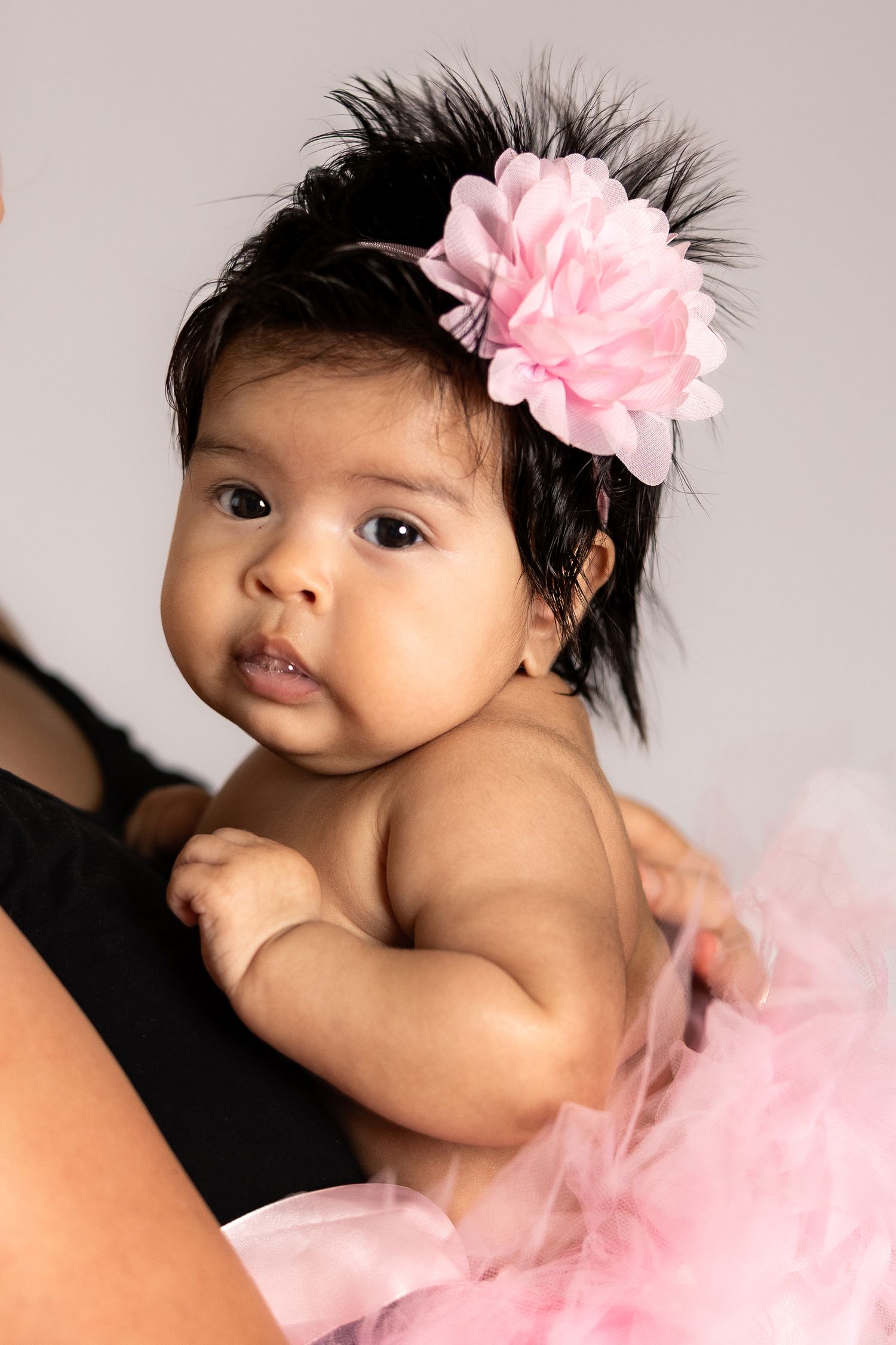 Adorable baby girl wearing a pink tutu and floral headband with feathers, held by an adult in a studio portrait.