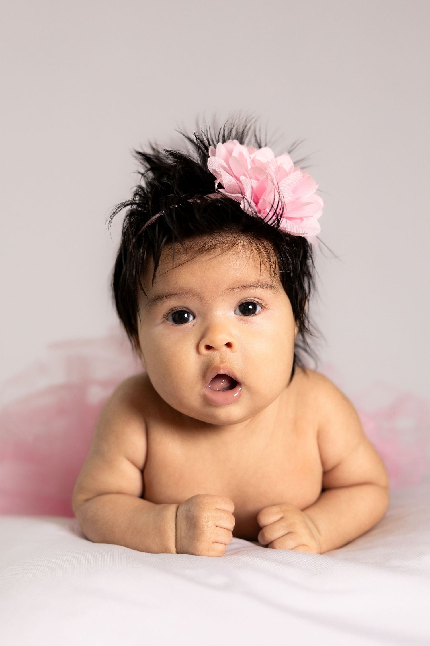 Adorable baby girl with dark hair wearing a pink flower headband, lying on her tummy with a surprised expression.