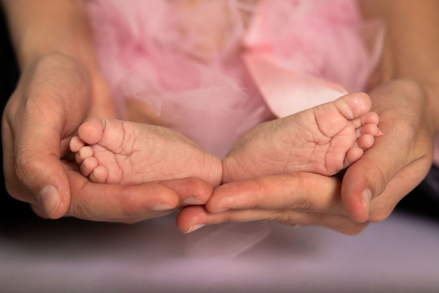 Newborn baby tiny feet cradled gently in adult hands, wearing pink outfit, close-up photography.