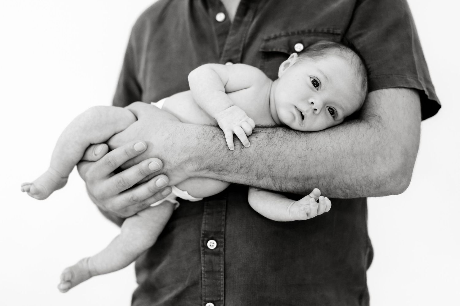 Black and white photo of a father cradling his newborn baby in one arm against a white background.