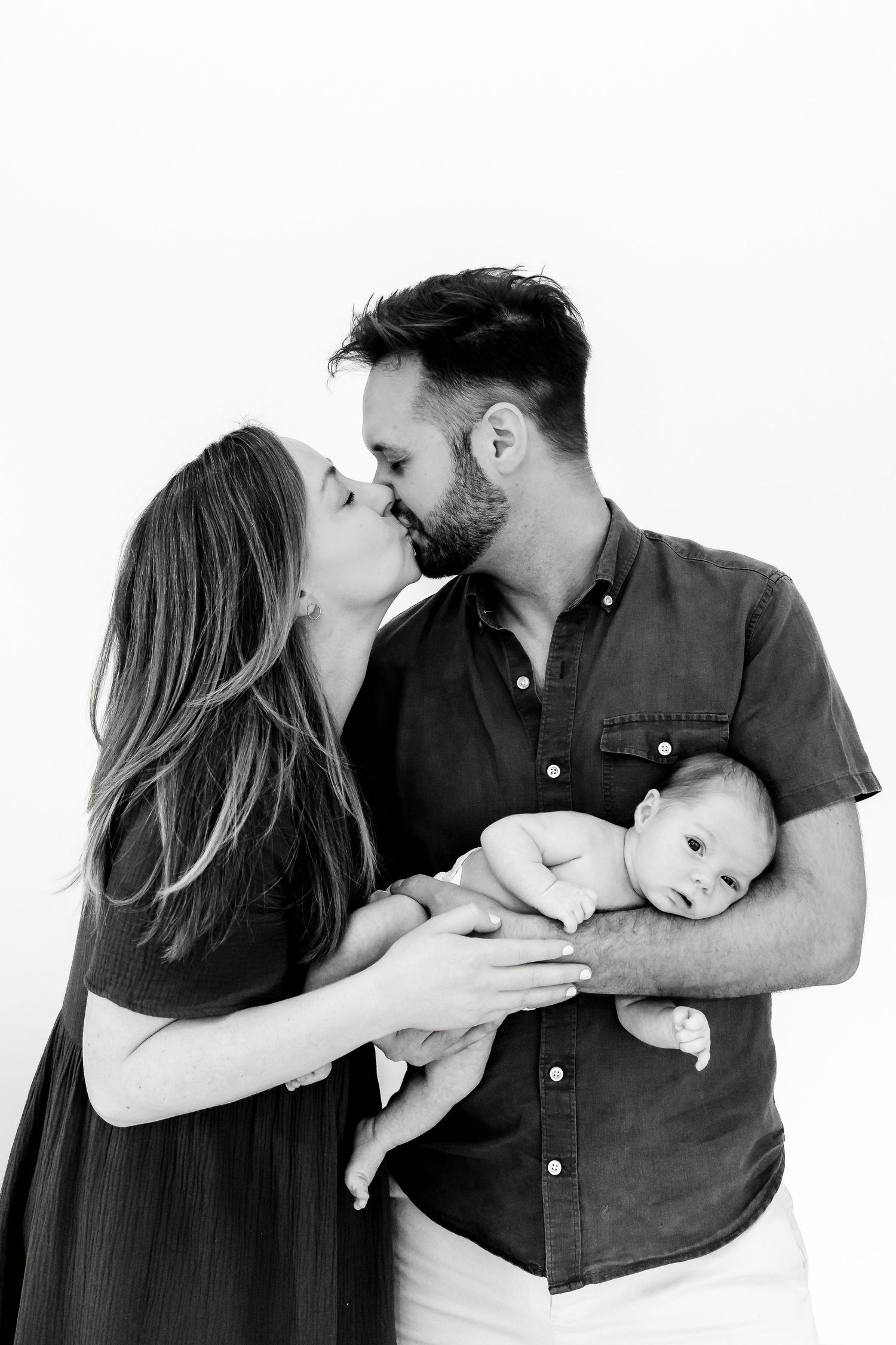 Black and white photo of a couple kissing while tenderly holding their newborn baby against a white background.