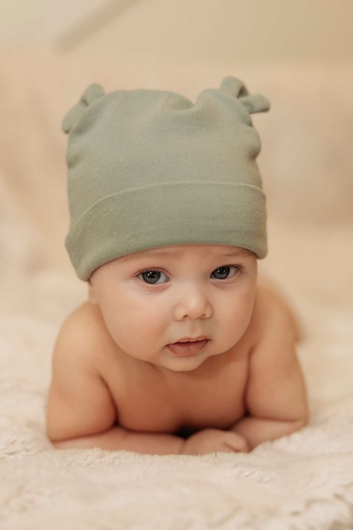 Adorable baby wearing a sage green knot hat lying on tummy on a soft cream blanket, looking at camera.