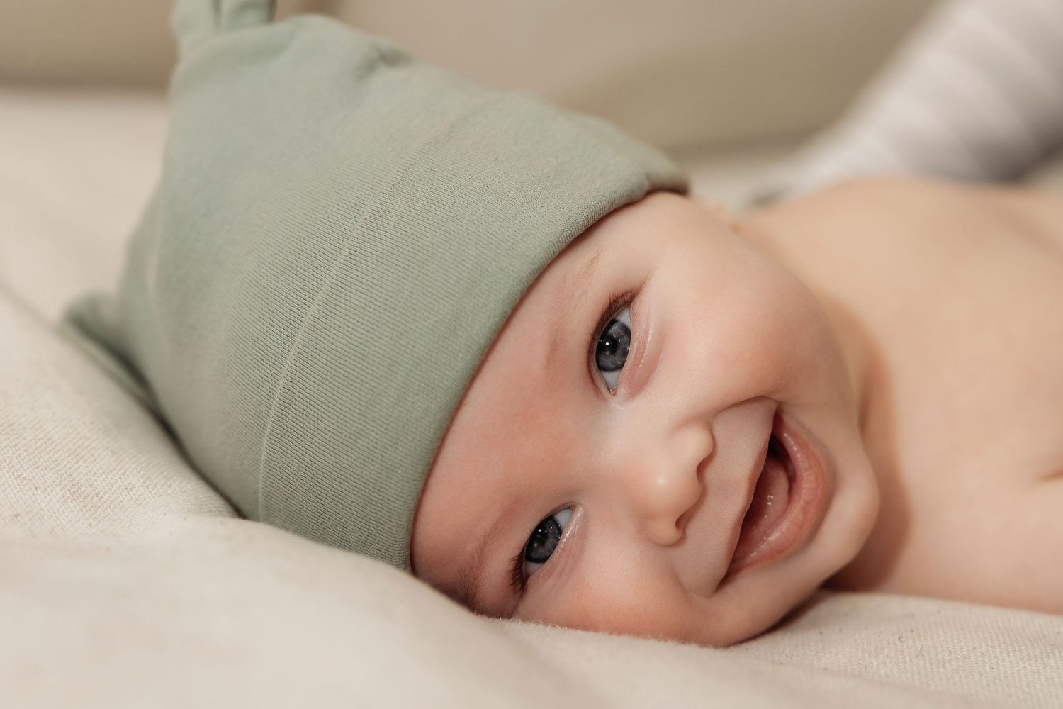 Smiling newborn baby wearing a sage green knit hat lying on a soft white blanket with bright blue eyes.