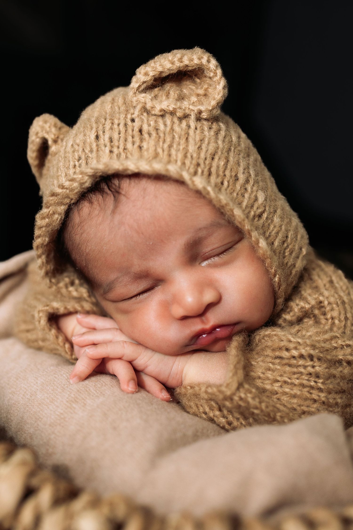 Sleeping newborn baby wrapped in beige knit bonnet on soft neutral backdrop.