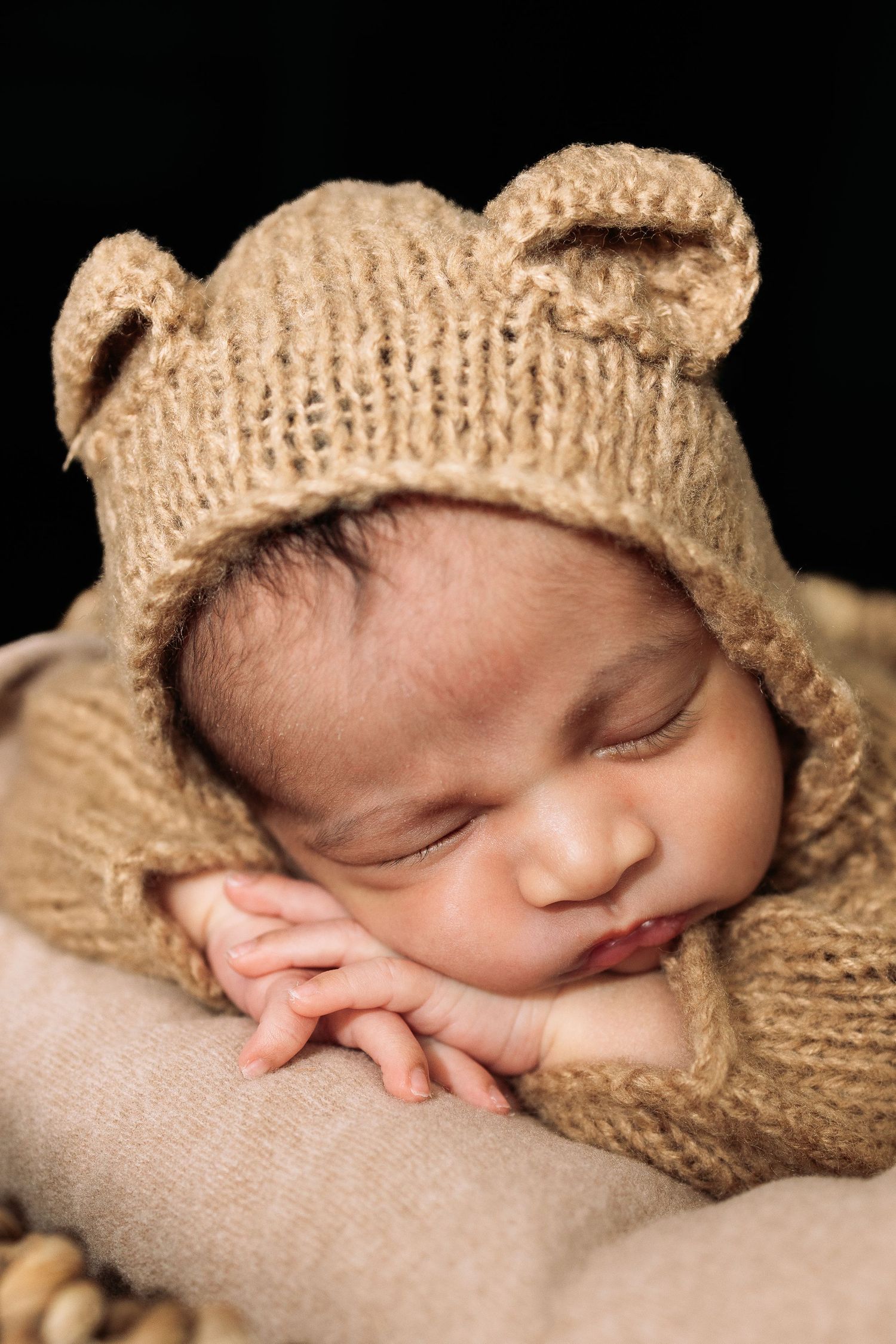Peaceful newborn sleeping in tan knitted wrap against dark background.