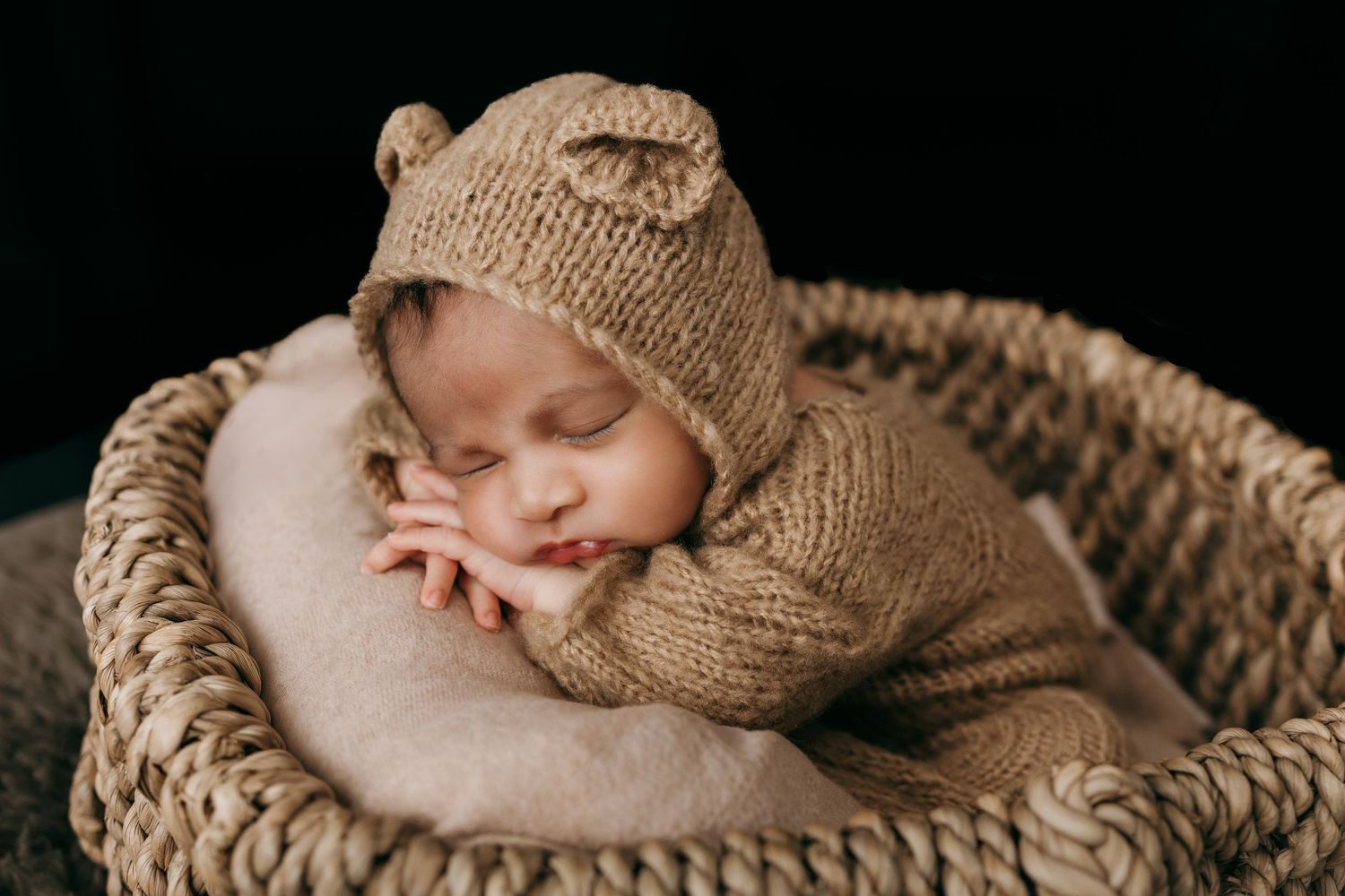 Sleeping newborn wrapped in beige knit blanket on dark backdrop.