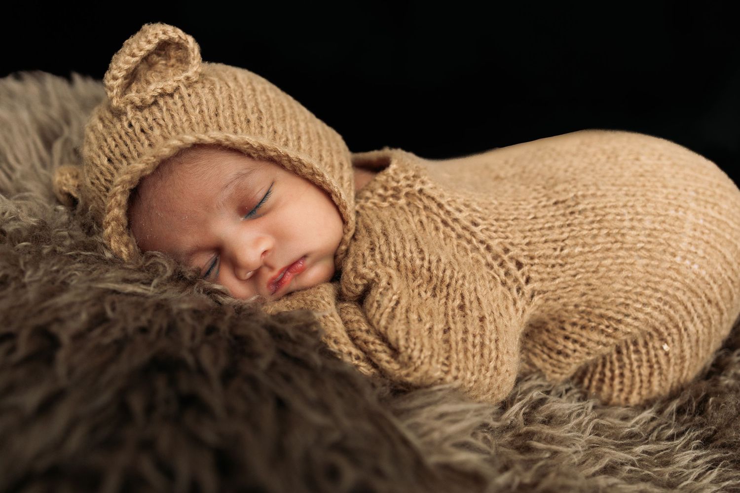 Baby in soft knitted beige clothing sleeping on textured surface.