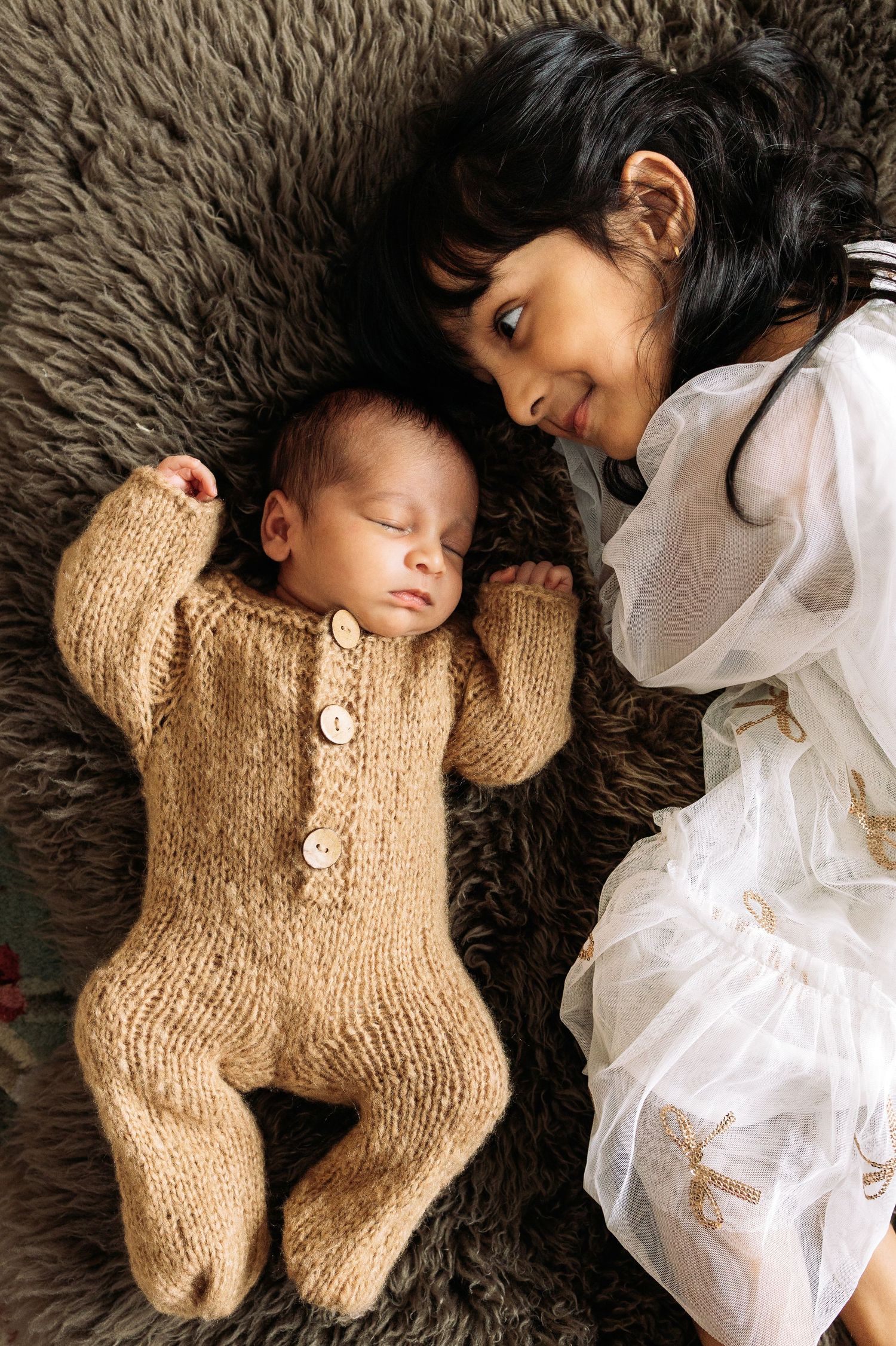 Baby in knit onesie sleeping on furry blanket next to sibling in white dress.