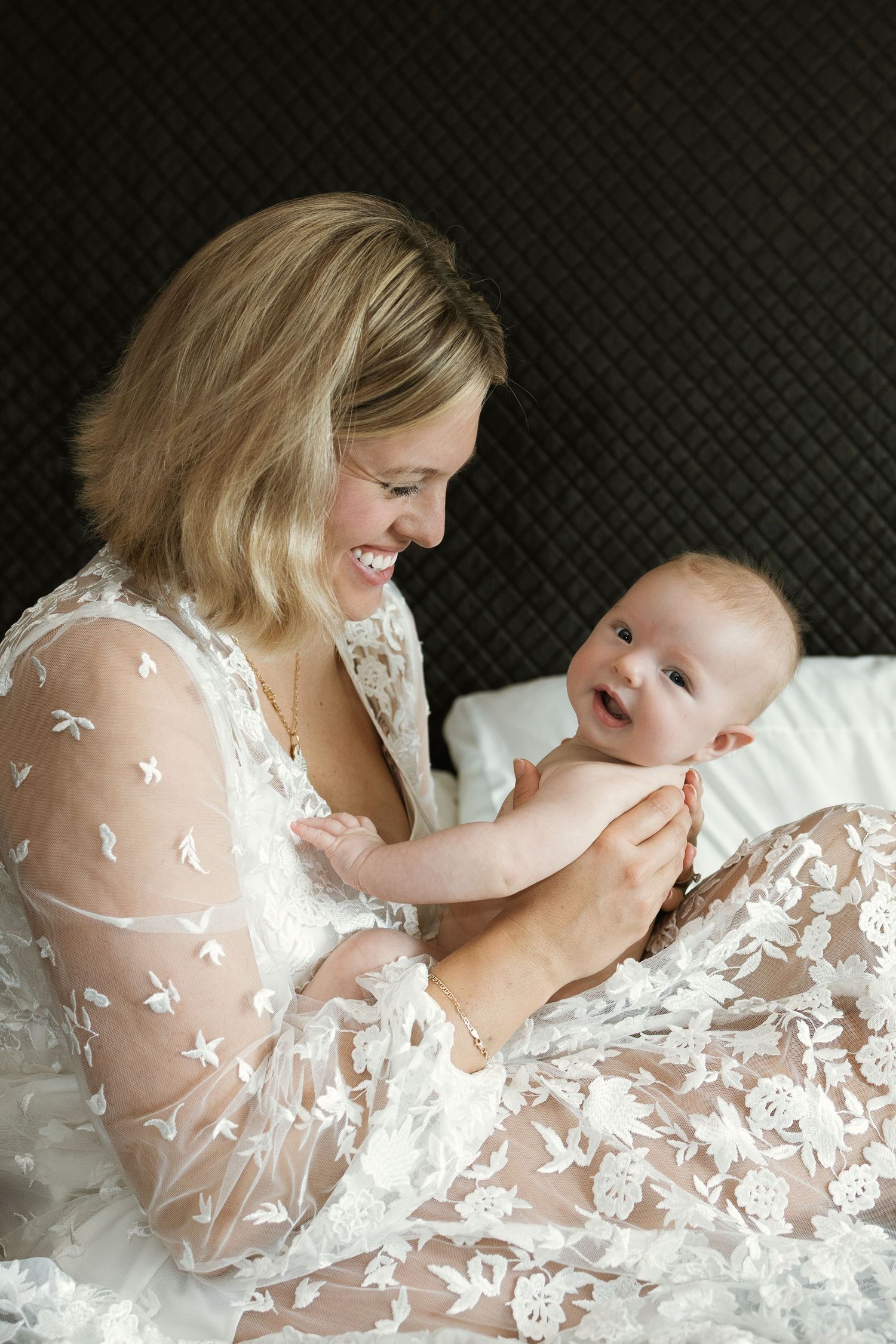 Mother in delicate lace gown sharing intimate moments with baby on white bedding.