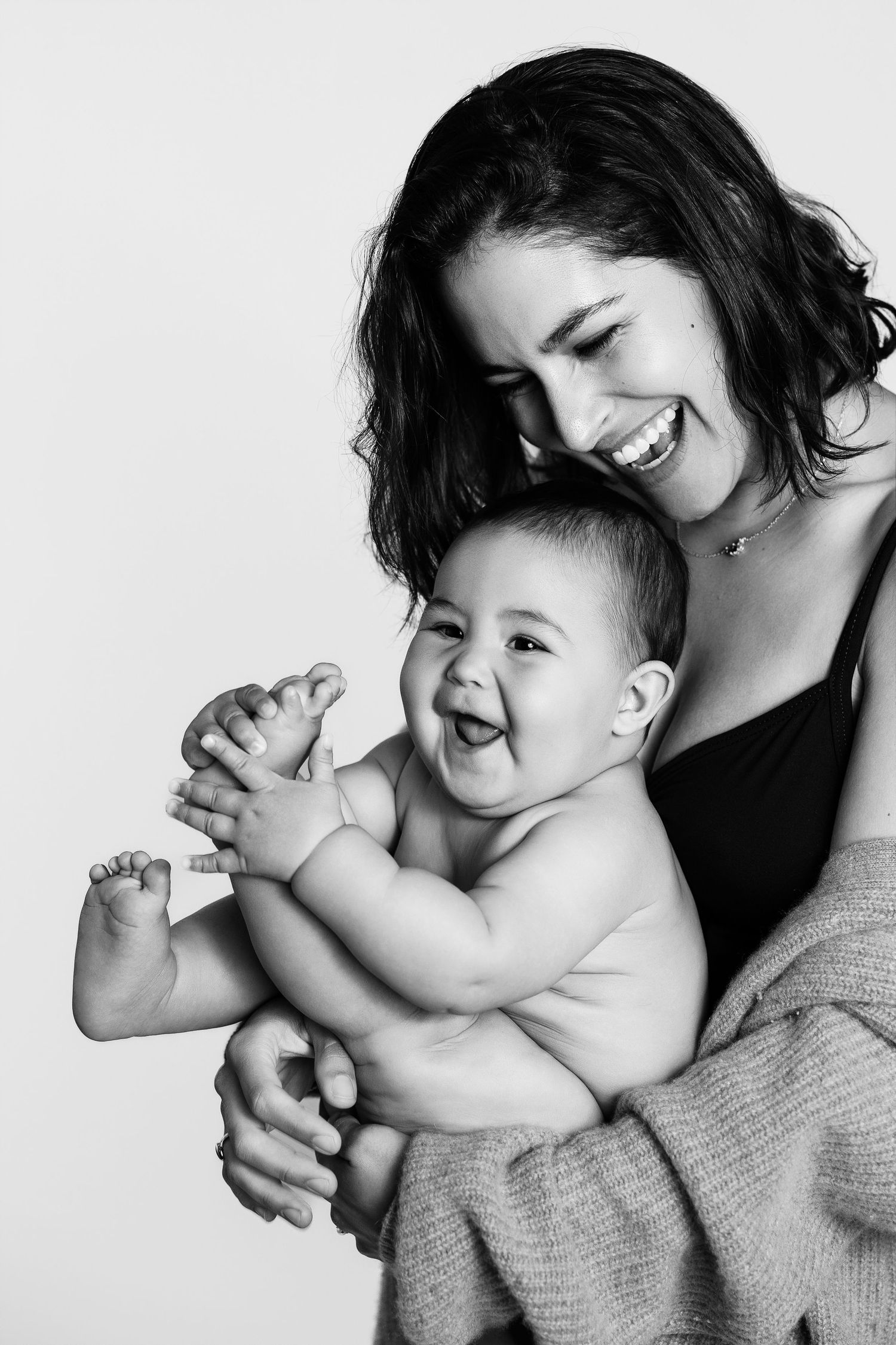 Black and white photo of a laughing mother holding her happy, giggling baby against a light background.