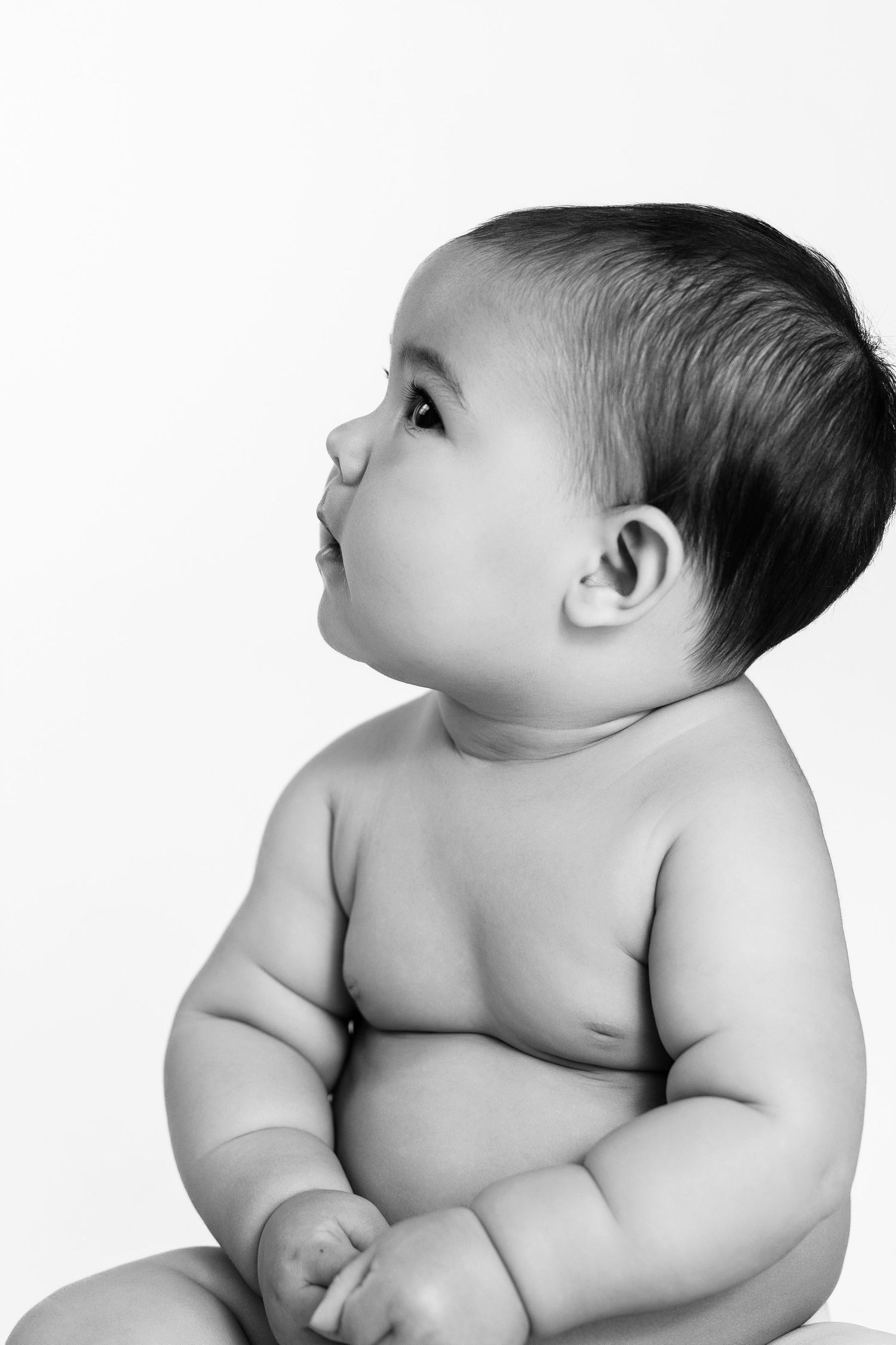 Black and white portrait of a cute chubby baby sitting upright, looking up curiously against a white background.