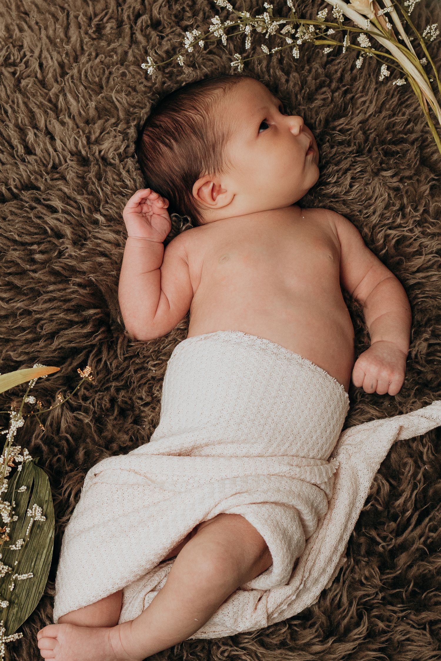 Newborn baby wrapped in white cloth lying on brown textured blanket with dried flowers.
