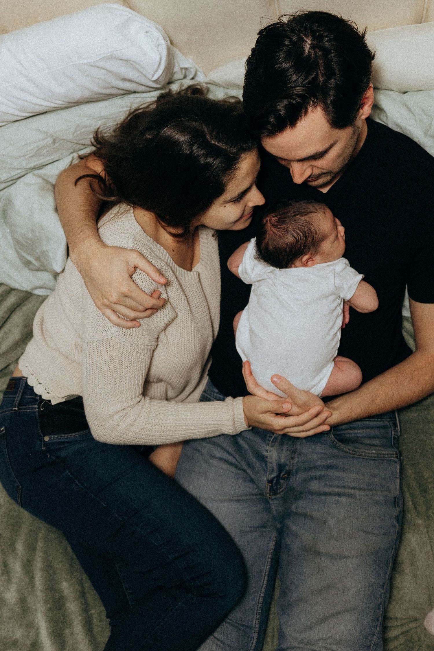 Intimate family portrait session showing parents cuddling with newborn on bed.
