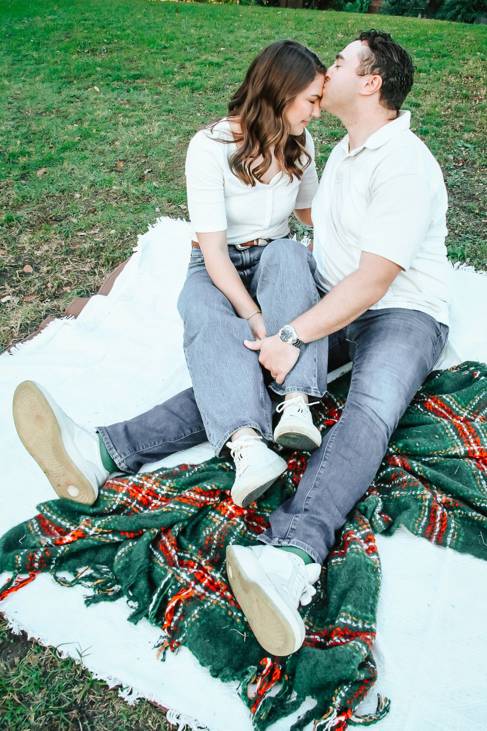 Couple sharing a gentle kiss while sitting on a plaid blanket in a grassy park.