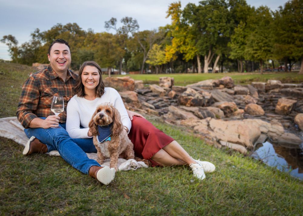 Smiling couple sitting outdoors with their golden doodle dog, holding wine glasses near a rocky stream.