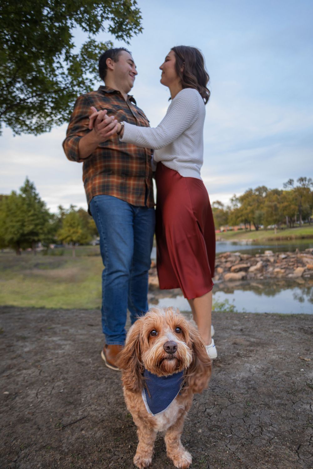 Couple dancing and laughing outdoors near a lake with an adorable fluffy golden dog wearing a blue bandana in the foreground.
