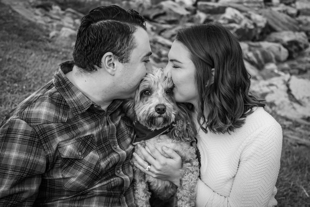 Black and white photo of couple kissing their fluffy dog on the cheeks outdoors.
