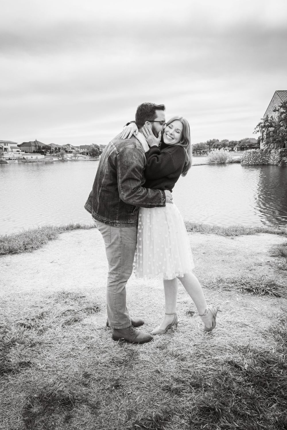 Black and white photo of a couple sharing a tender kiss while embracing near a peaceful lakeside setting.