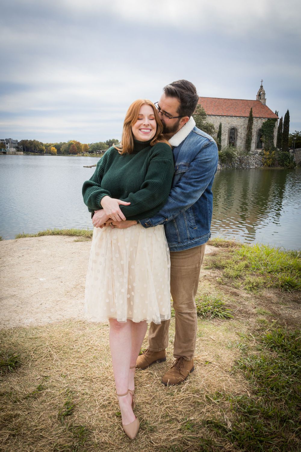 Couple embracing and smiling together by a lake with a charming stone chapel visible in the background.