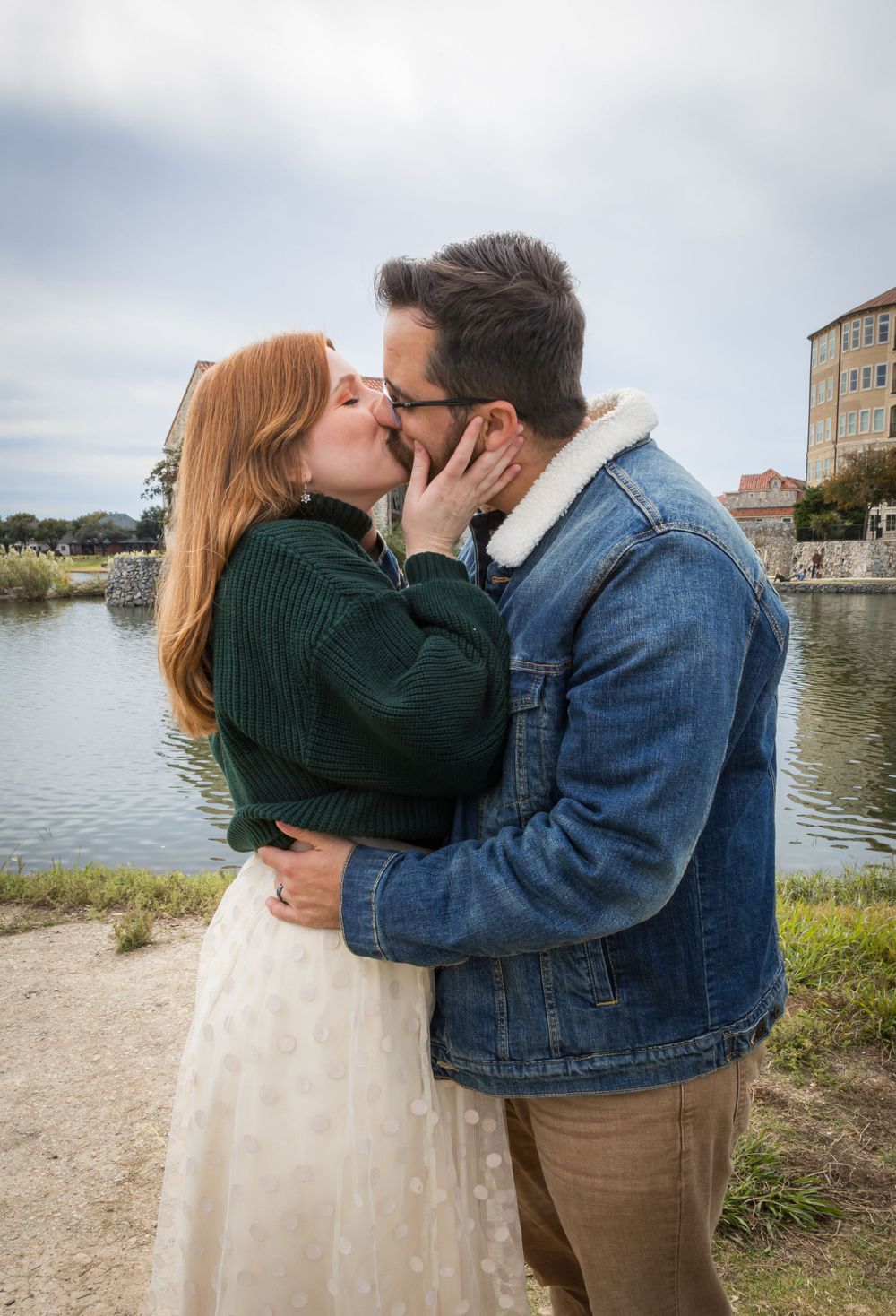 Romantic couple sharing a passionate kiss by a lakeside with a historic stone building in the background.